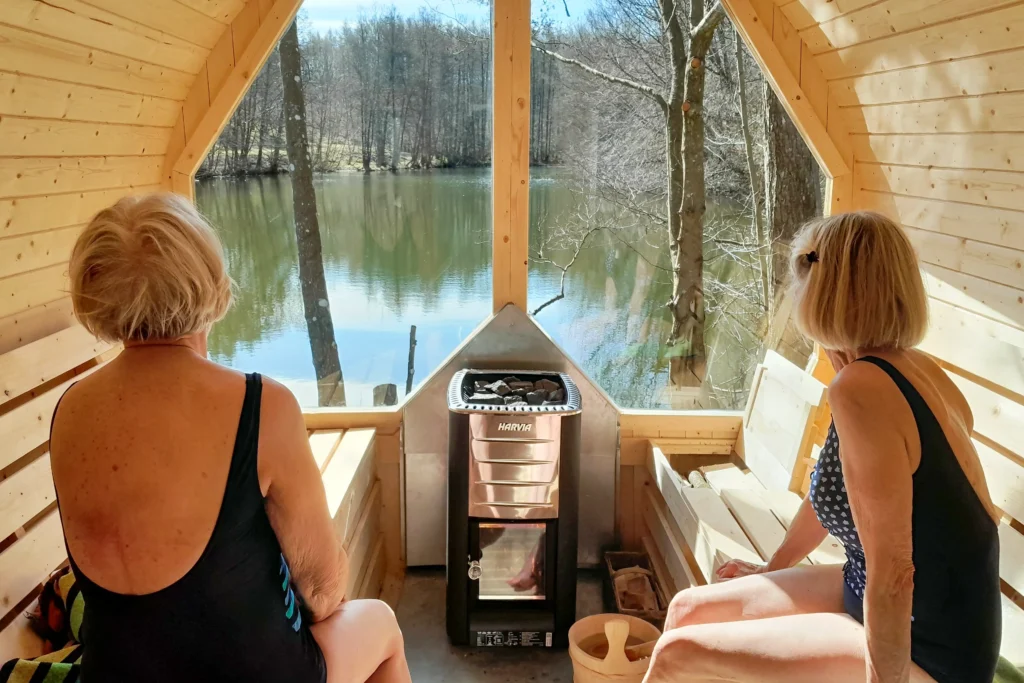Two older women sitting in a wood-fired sauna looking out a big window at a small lake.