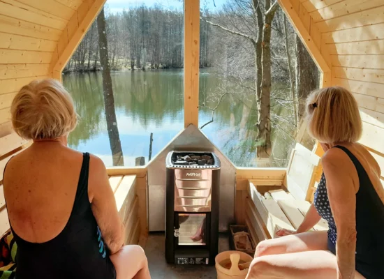 Behind the wood-stove there is a big window facing the small lake. Two older women sitting in a wood-fired sauna looking out a big window at a small lake.